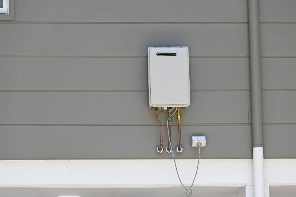 Wall-mounted tankless water heater with exposed copper plumbing and shutoff valves installed on the exterior wall of a home.