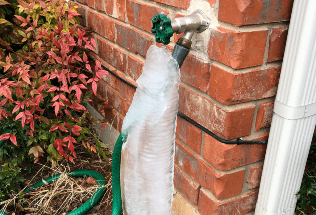 A frozen outdoor faucet is covered in thick ice, with a green garden hose attached beside a brick exterior wall.