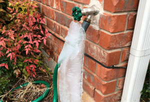 A frozen outdoor faucet is covered in thick ice, with a green garden hose attached beside a brick exterior wall.